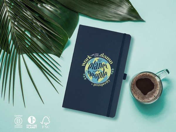Notebook, journal on light blue background with palm leaves and coffee. Dark blue journal with a graphic that has a planet and the words Work Like You Give a Damn about People, Planet and Prosperity.